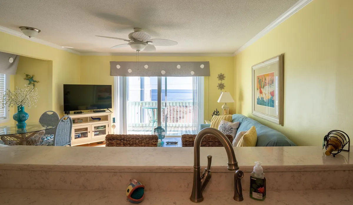 A cozy living area with a comfortable sofa facing a television, and a view extending out to a balcony through sliding glass doors. Soft daylight fills the room, and ocean views can be glimpsed from the seating area, reinforcing the seamless connection between indoor relaxation and outdoor scenery.