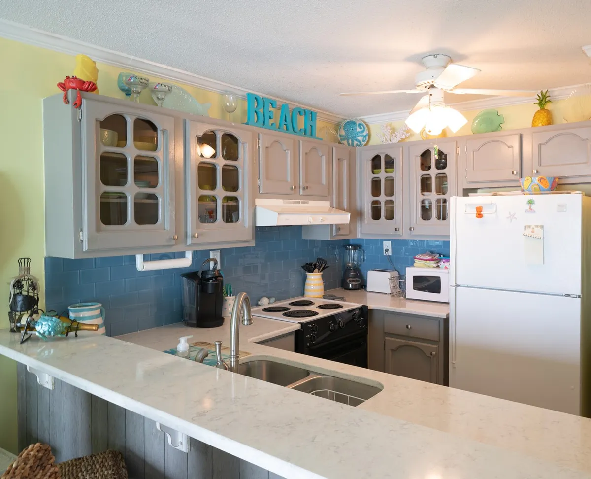 A bright, beach-themed kitchen with colorful cabinets, a marble countertop, and modern appliances. Large windows allow natural light to flood in, and the playful decor evokes a cheerful coastal vibe. The kitchen is adjacent to a breakfast nook, ideal for enjoying morning coffee with a view.
