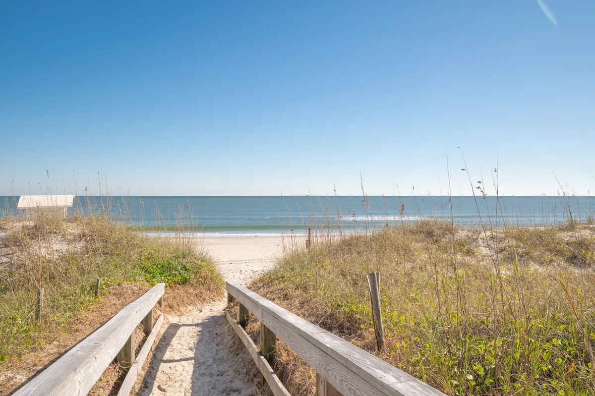 A tranquil view of a wooden boardwalk stretching over sandy dunes toward the ocean, with the beach and sky beyond. The scene captures the anticipation of reaching the shore, emphasizing the coastal setting.