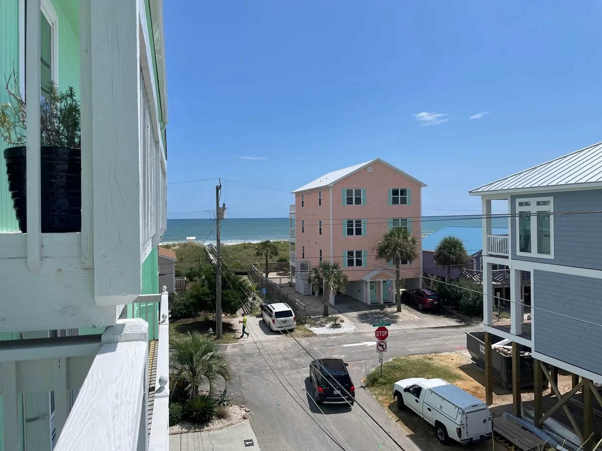 Overlooking the ocean, this image features a balcony with a green plant, colorful houses, and a panoramic sea view. The perspective invites viewers to imagine relaxing outdoors, savoring a sunrise, or enjoying quiet time with a book while soaking in the coastal atmosphere.