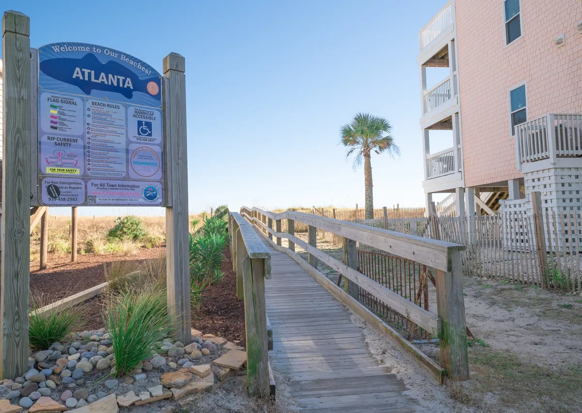 A cheerful exterior scene featuring a prominent 'Welcome' sign, a wooden walkway leading towards the beach, and a palm tree beside the path. The view offers a clear sense of arrival and direct access to the sand and surf.