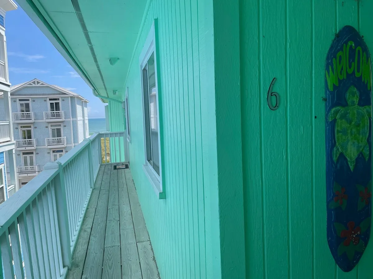 The image features the exterior corridor of the condo community, with a welcoming door marked '6' and a cheerful turtle-themed welcome sign. The corridor opens up to the coastal surroundings, suggesting proximity to the ocean and communal spaces.