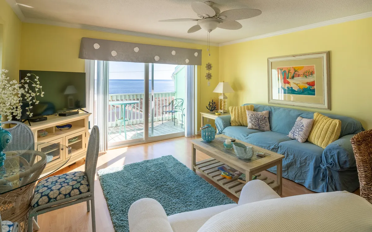 This inviting living room features a plush sofa and coffee table, set beside large balcony doors that open to sunrise ocean views. The space is bathed in cheerful natural light, perfect for relaxing, entertaining, or enjoying quiet mornings overlooking Carolina Beach.
