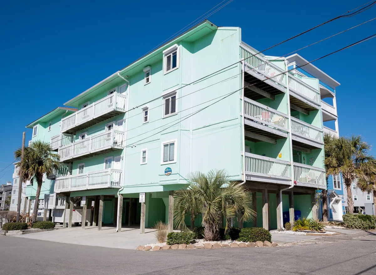 An exterior shot of a brightly colored building with palm trees and balconies, set against a sunny sky. The scene hints at nearby beach access and the vibrant coastal ambiance that defines the area, with suggestions of outdoor gatherings and local seafood restaurants close by.