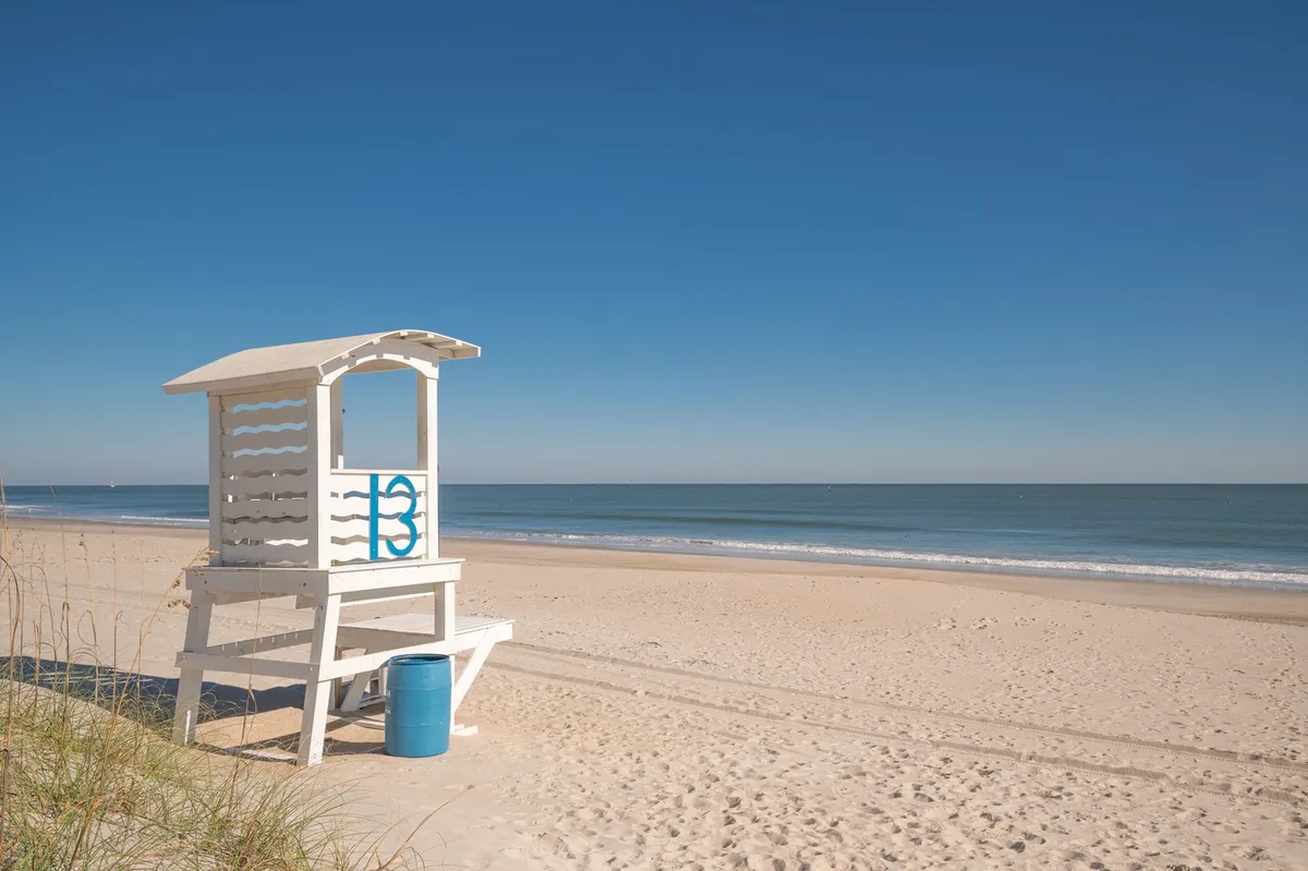 A lively beach scene with a classic lifeguard tower, blue barrel, sandy shore, and ocean waves. The area is inviting for sunbathing, swimming, and enjoying family beach activities.