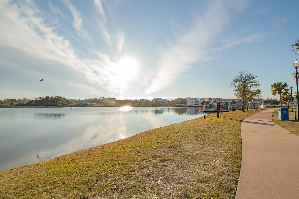 A serene lakeside scene featuring a winding pathway, calm water, sun filtering through trees, and buildings in the background. The space invites leisurely strolls, picnics, and peaceful relaxation in a natural setting.
