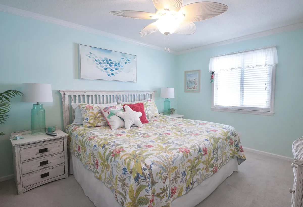 A tranquil bedroom scene with a floral-patterned bed, beach-themed artwork, and a cozy nightstand with a lamp. The room is filled with natural light, enhancing the cheerful and inviting atmosphere, making it perfect for guests or family members.