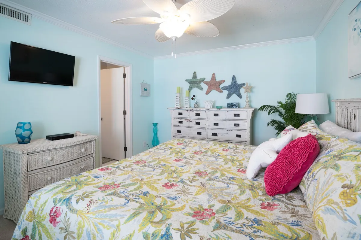 A cozy bedroom with a colorful bedspread, decorative starfish, a tropical plant, wall-mounted TV, and a spacious dresser. Ample sunlight streams in, offering a peaceful retreat for rest or relaxation.
